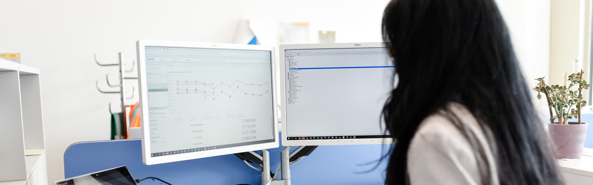 Eine Mitarbeiterin steht mit dem Rücken zur Kamera an ihrem Arbeitsplatz und schaut in ihren PC Monitor rein.