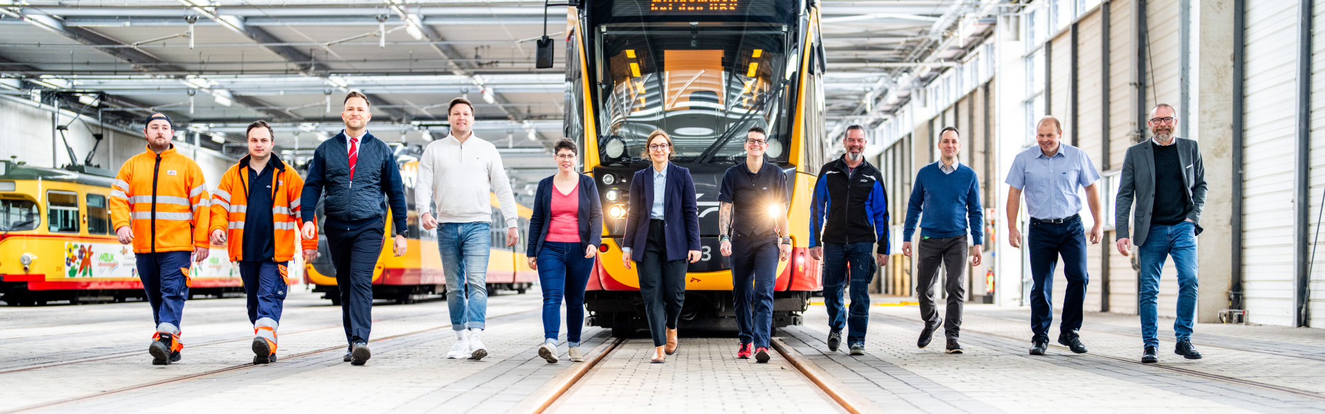 Mitarbeitende verschiedener Berufsbereiche laufen vor einer Bahn in der Bahnhalle.