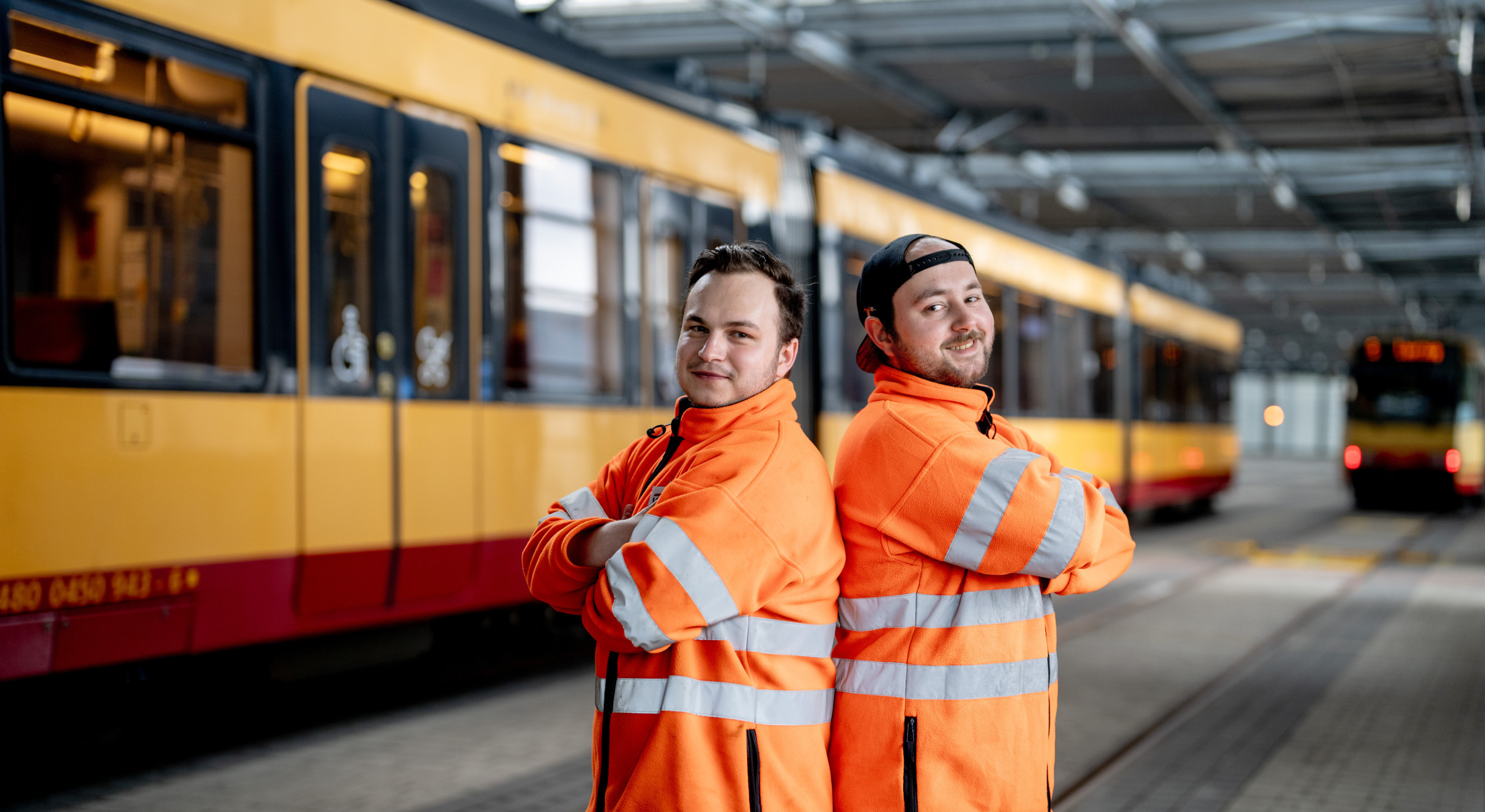 Zwei junge Mitarbeitende stehen lächelnd in orangenen Arbeitsjacken Schulter an Schulter. Im Hintergrund ist eine Bahn zu sehen.