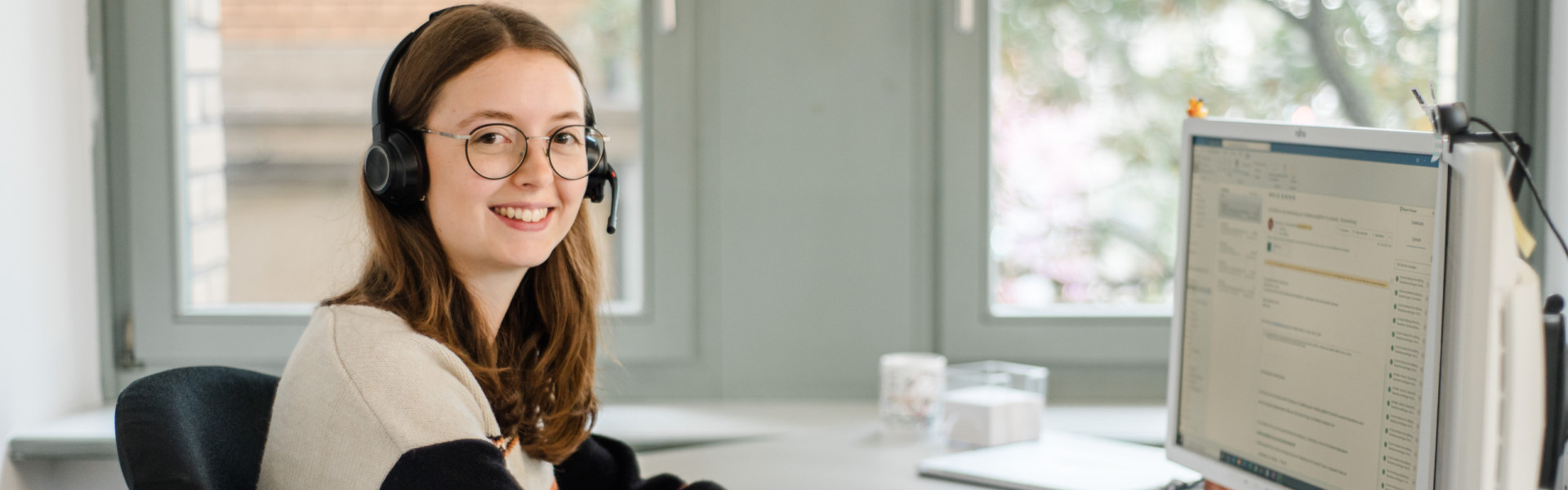 Eine Mitarbeiterin sitz mit Headset an einem Schreibtisch und bearbeitet die Anliegen der Kunden. Im Hintergrund scheint durch zwei Fenster viel Tageslicht.