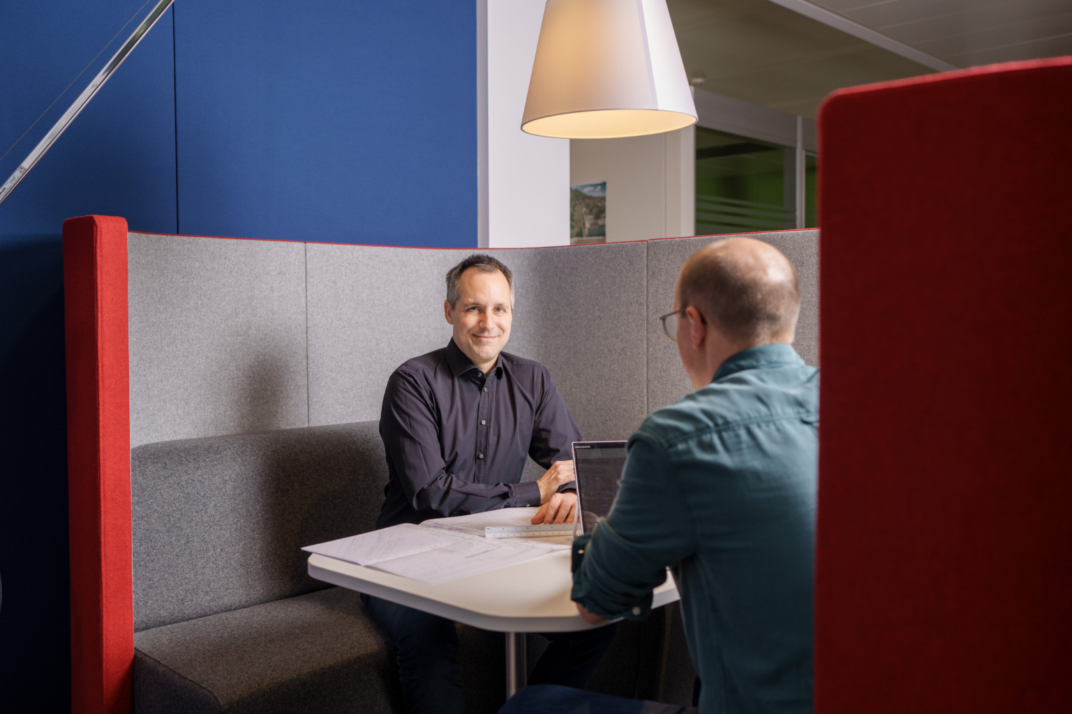 Zwei Mitarbeiter sitzen in einer Kabine wobei der eine mit dem Rücken zur Kamera sitzt und der andere in die Kamera hineinschaut.