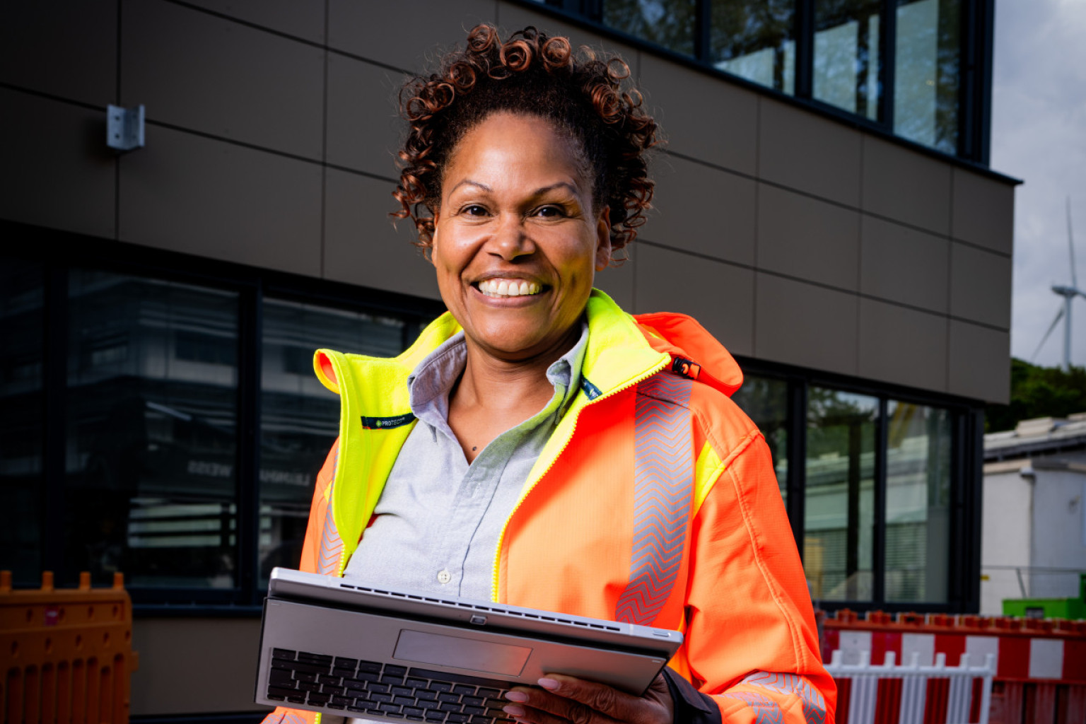 Die Projektplanerin steht mit einem Helm in der einer und einem Tablet in der anderen Hand und steht vor einem Gebäude.