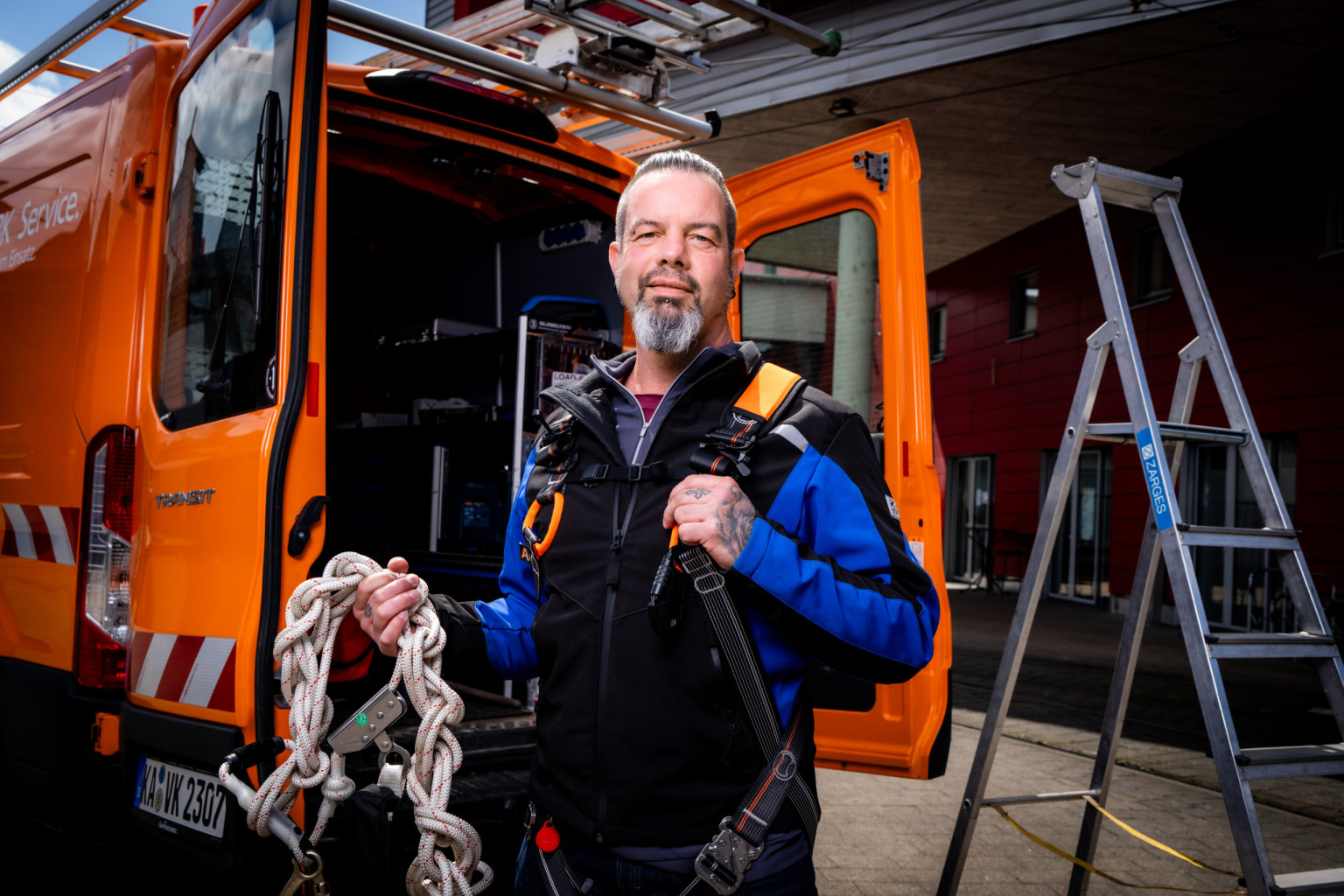 Ein Mitarbeiter steht mit Sicherheitsgurt und Seil vor einem Unternehmensfahrzeug.